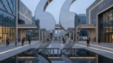 Large reflective sculpture shaped like two curved quotation marks in a modern plaza, mirrored in a shallow pool with surrounding buildings and pedestrians.